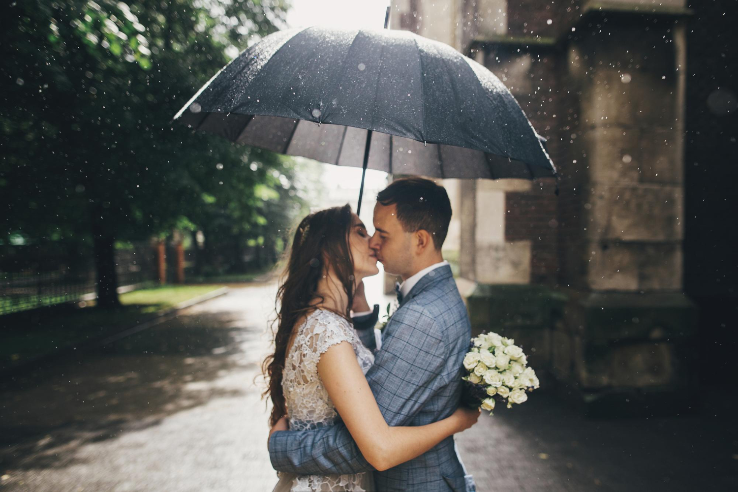 stylish bride and groom kissing under umbrella on background of old church in rain